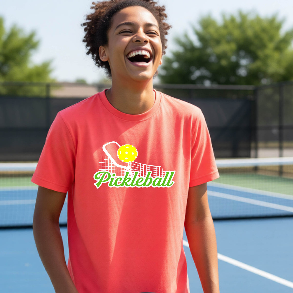 A young person laughing on an outdoor pickleball court wearing a bright coral pink t-shirt with a yellow and white pickleball graphic and the word 'Pickleball' printed on the front.