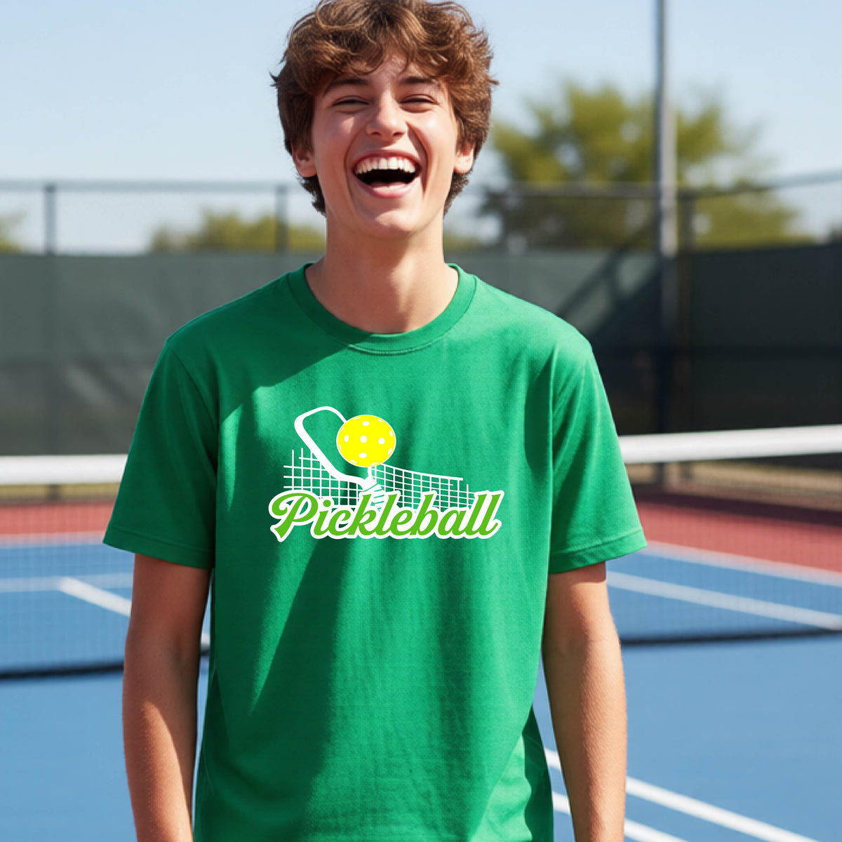 A young person laughing on an outdoor pickleball court wearing a bright green t-shirt with a yellow and white pickleball graphic and the word 'Pickleball' printed on the front.
