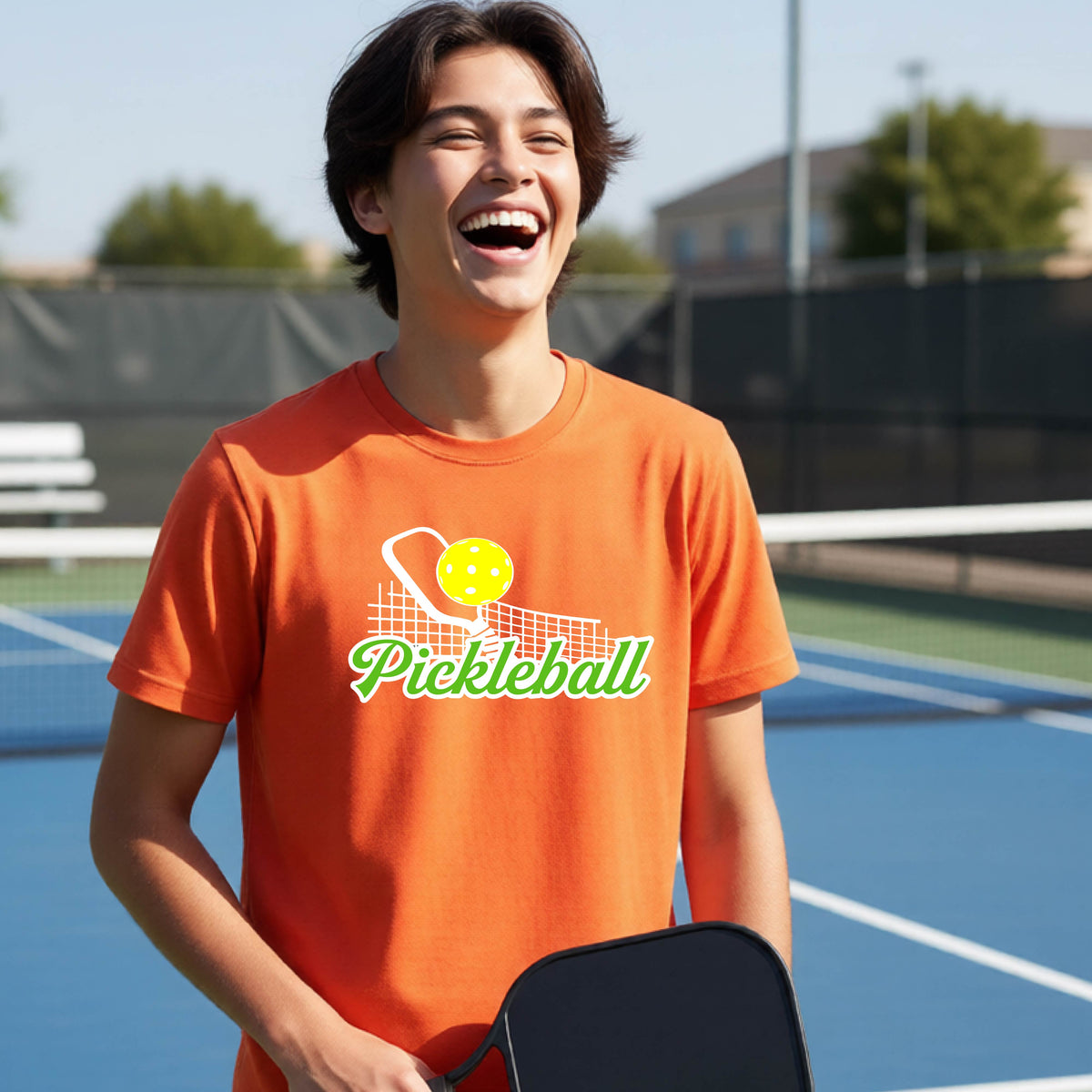 A young person laughing on an outdoor pickleball court wearing a bright orange t-shirt with a yellow and white pickleball graphic and the word 'Pickleball' printed on the front.