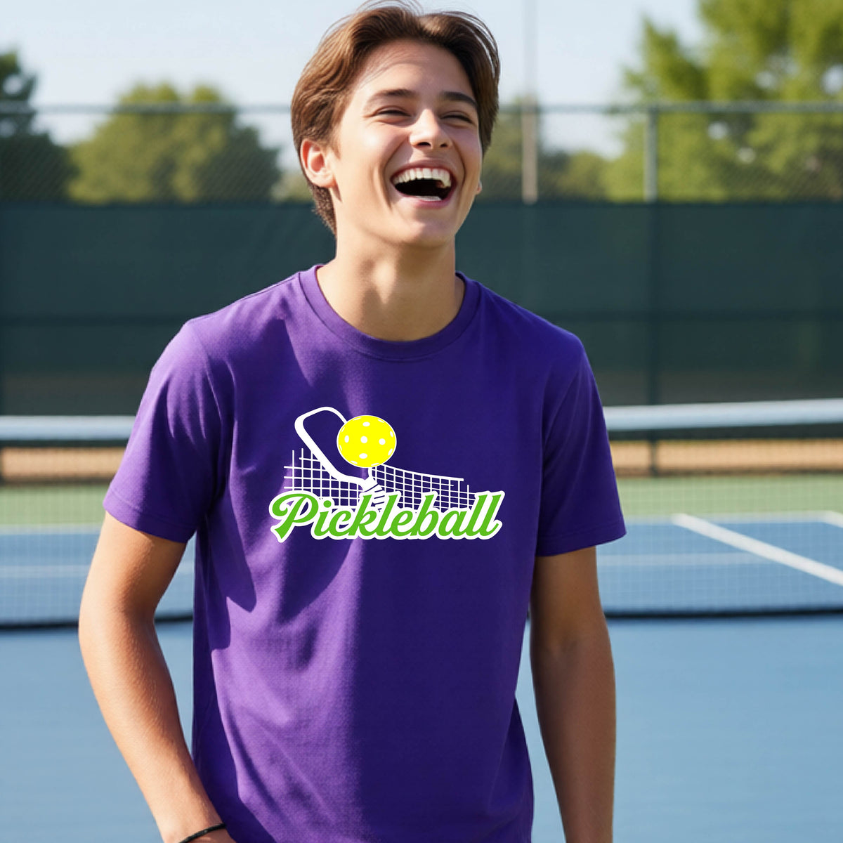 A young person laughing on an outdoor pickleball court wearing a purple t-shirt with a yellow and white pickleball graphic and the word 'Pickleball' printed on the front.