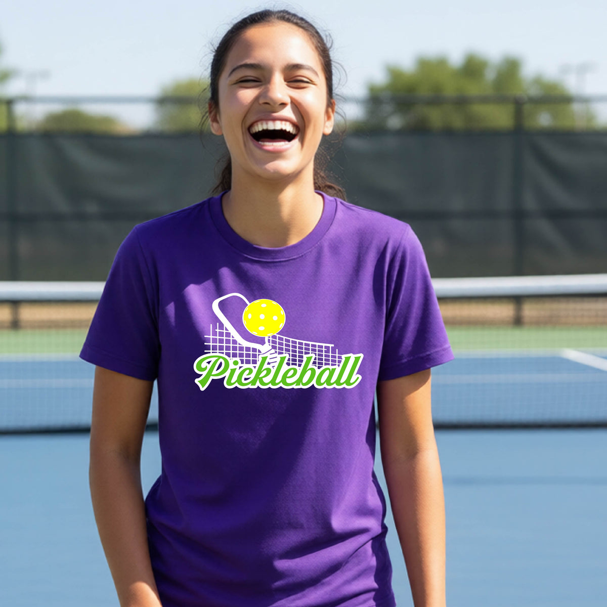 A young person laughing on an outdoor pickleball court wearing a purple t-shirt with a yellow and white pickleball graphic and the word 'Pickleball' printed on the front.