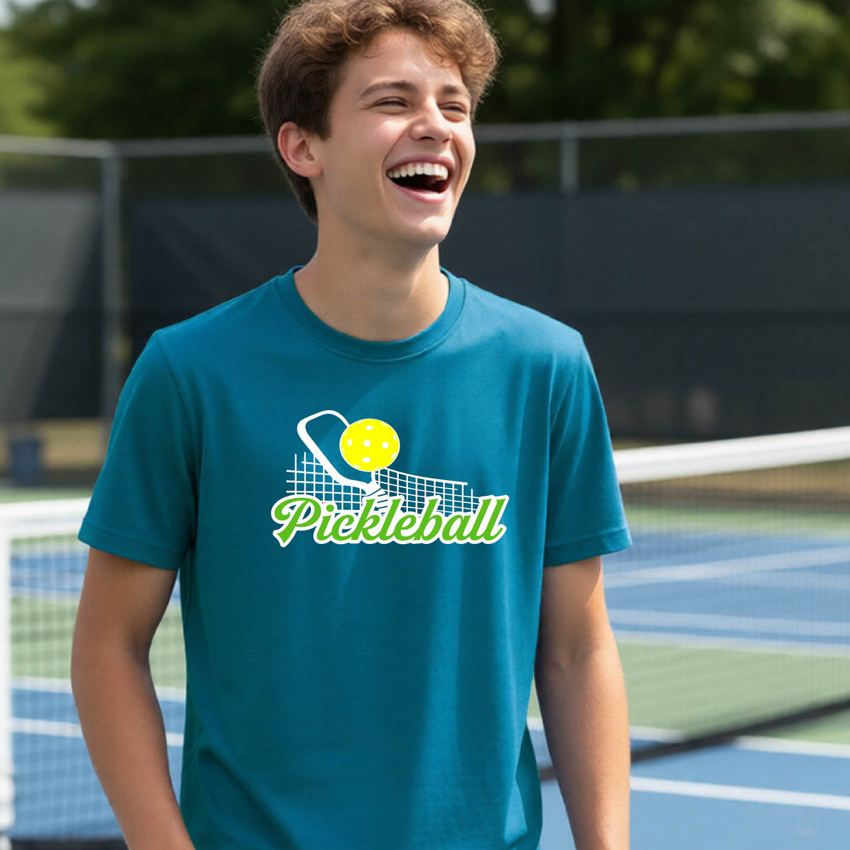 A young person laughing on an outdoor pickleball court wearing a teal t-shirt with a yellow and white pickleball graphic and the word 'Pickleball' printed on the front.