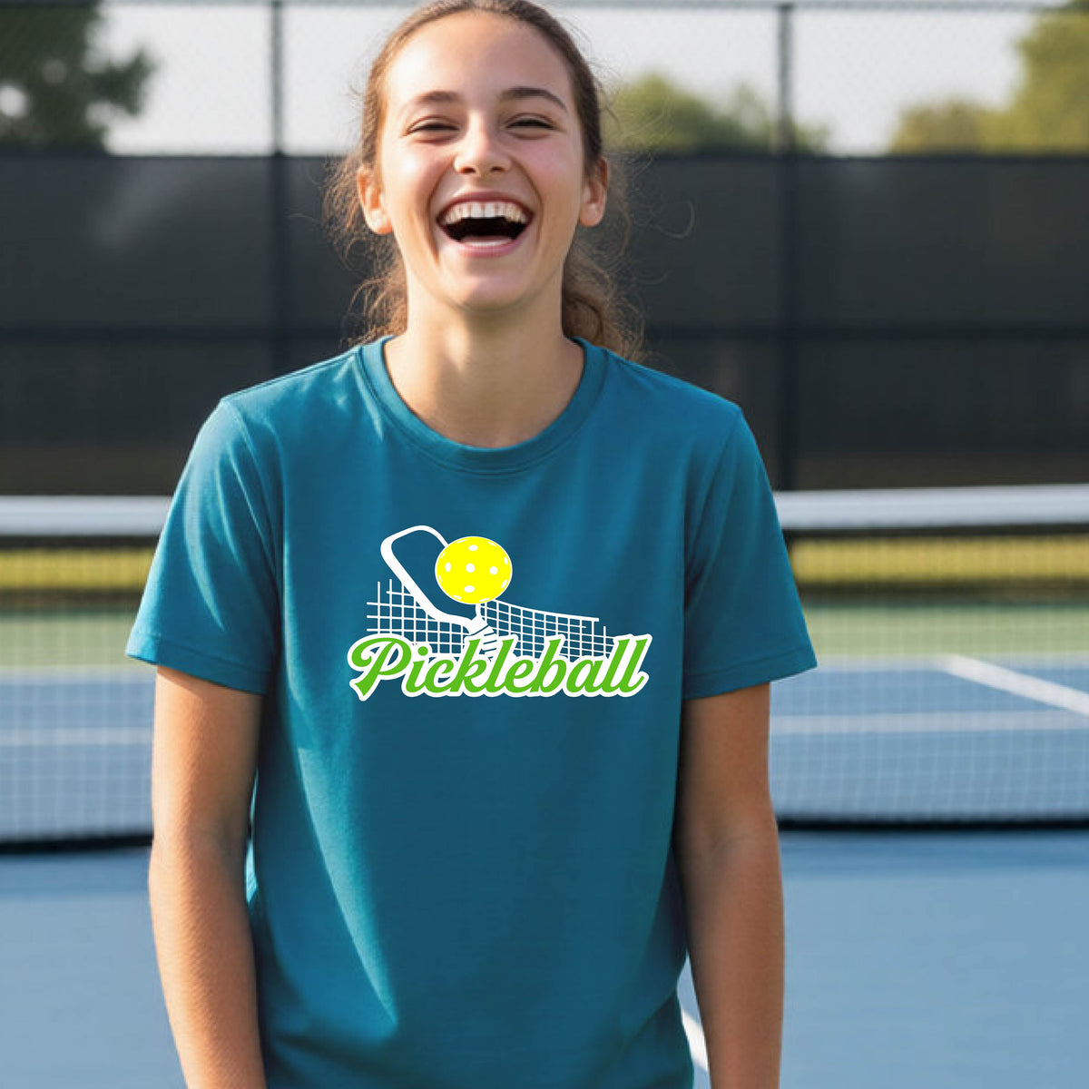 A young person laughing on an outdoor pickleball court wearing a teal t-shirt with a yellow and white pickleball graphic and the word 'Pickleball' printed on the front.