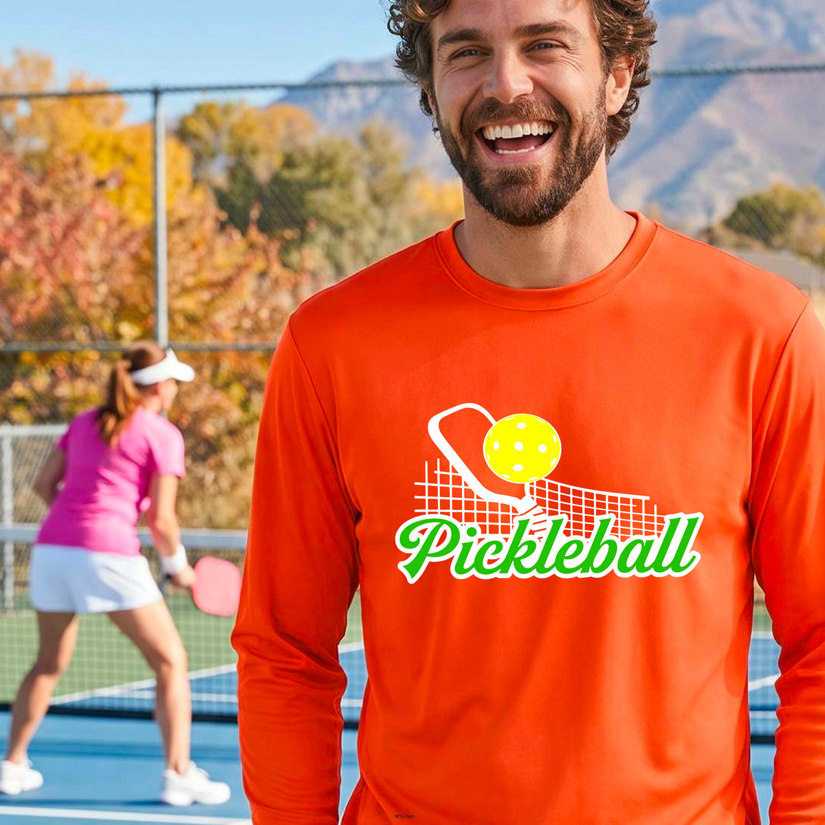 A woman wearing an orange long-sleeve t-shirt with a graphic on the front that has white and green text "PICKLEBALL'  with a white pickleball paddle and net and a yellow pickleball above the text graphic.