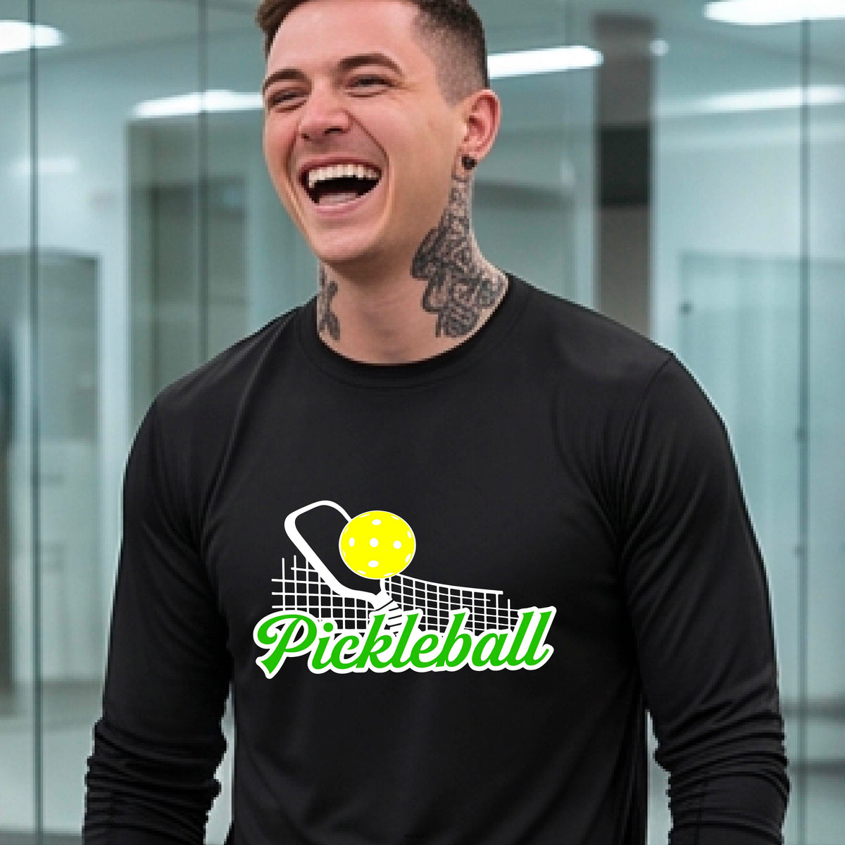 A woman wearing a black long-sleeve t-shirt with a graphic on the front that has white and green text "PICKLEBALL'  with a white pickleball paddle and net and a yellow pickleball above the text graphic.