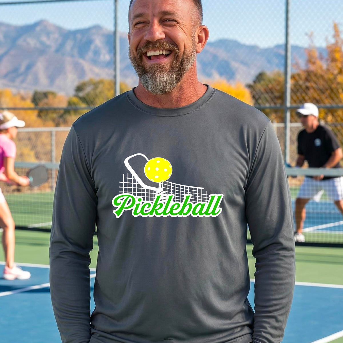 A woman wearing a gray long-sleeve t-shirt with a graphic on the front that has white and green text "PICKLEBALL'  with a white pickleball paddle and net and a yellow pickleball above the text graphic.