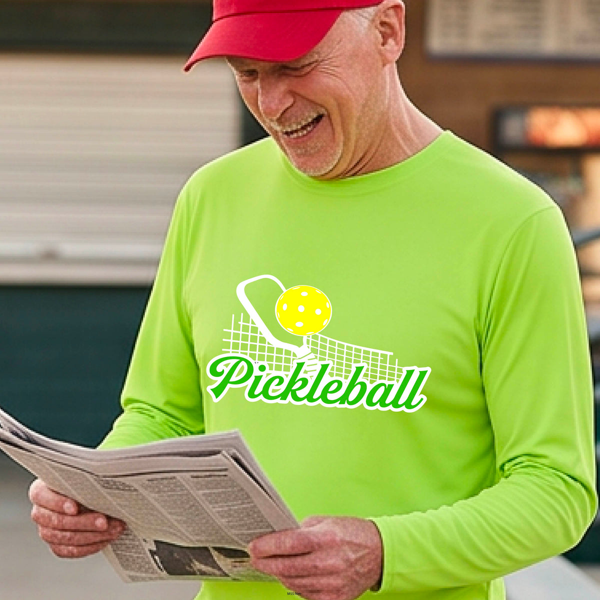 A woman wearing a lime green long-sleeve t-shirt with a graphic on the front that has white and green text "PICKLEBALL'  with a white pickleball paddle and net and a yellow pickleball above the text graphic.