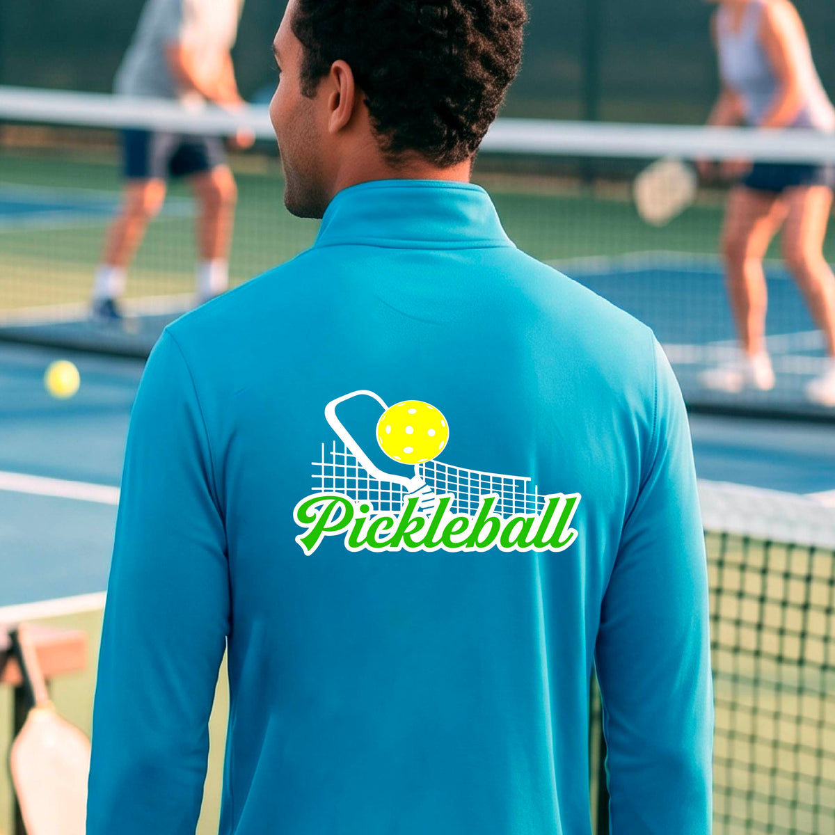 A close-up of a man wearing a bright blue long-sleeve quarter-zip shirt with a graphic on the front that has white and green text "PICKLEBALL'  with a white pickleball paddle and net and a yellow pickleball above the text graphic.