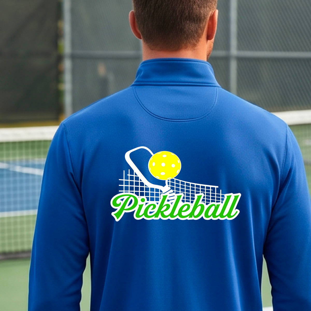 A close-up of a man wearing a royal blue long-sleeve quarter-zip shirt with a graphic on the front that has white and green text "PICKLEBALL'  with a white pickleball paddle and net and a yellow pickleball above the text graphic.