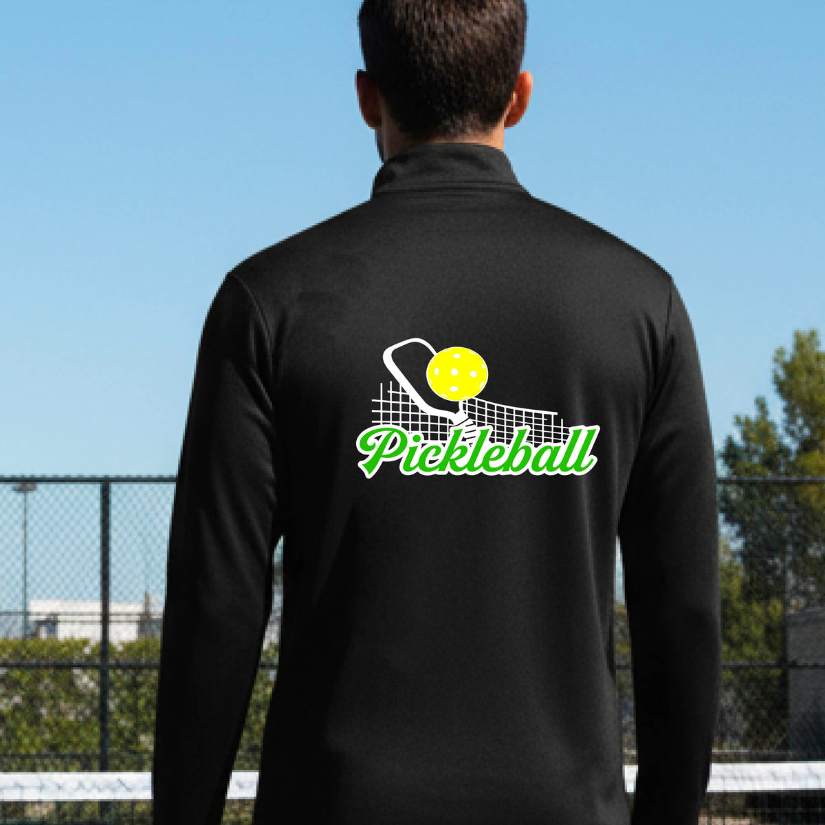 A close-up of a man wearing a black long-sleeve quarter-zip shirt with a graphic on the front that has white and green text "PICKLEBALL'  with a white pickleball paddle and net and a yellow pickleball above the text graphic.