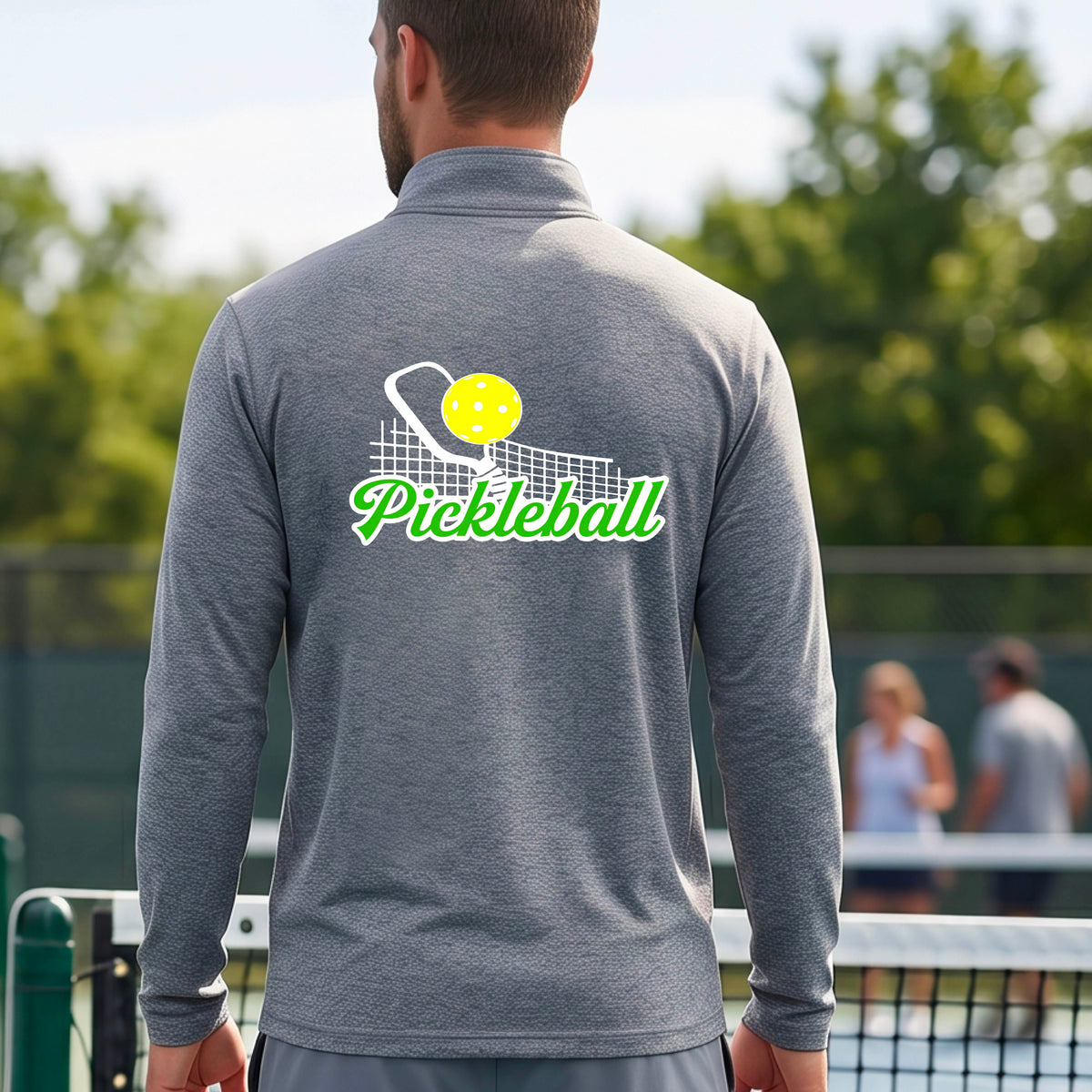 A close-up of a man wearing a gray long-sleeve quarter-zip shirt with a graphic on the front that has white and green text "PICKLEBALL'  with a white pickleball paddle and net and a yellow pickleball above the text graphic.