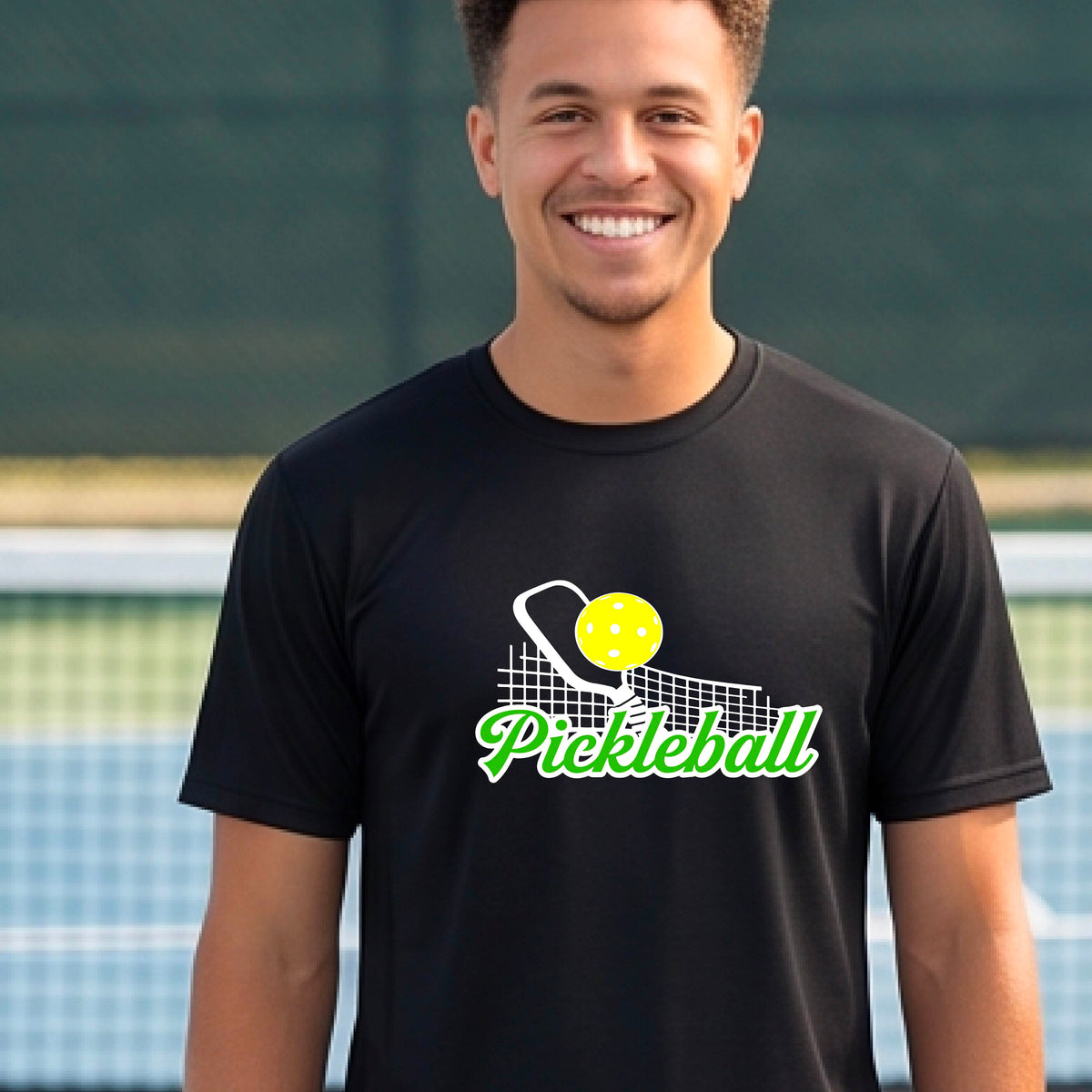 A man wearing a black short-sleeve t-shirt with a graphic on the front that has white and green text "PICKLEBALL'  with a white pickleball paddle and net and a yellow pickleball above the text graphic.