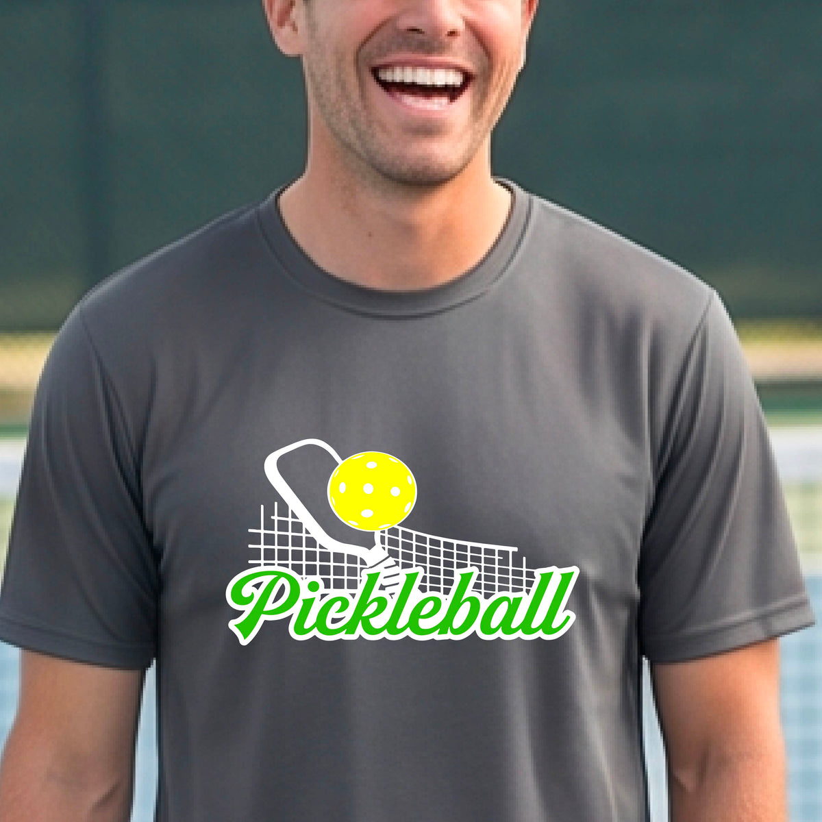 A man wearing a gray short-sleeve t-shirt with a graphic on the front that has white and green text "PICKLEBALL'  with a white pickleball paddle and net and a yellow pickleball above the text graphic.