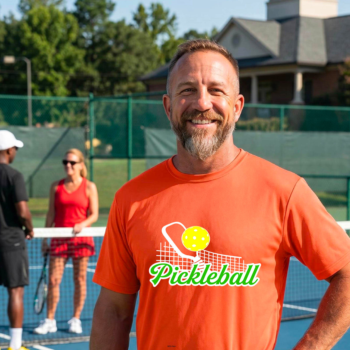 A man wearing an orange short-sleeve t-shirt with a graphic on the front that has white and green text "PICKLEBALL'  with a white pickleball paddle and net and a yellow pickleball above the text graphic.