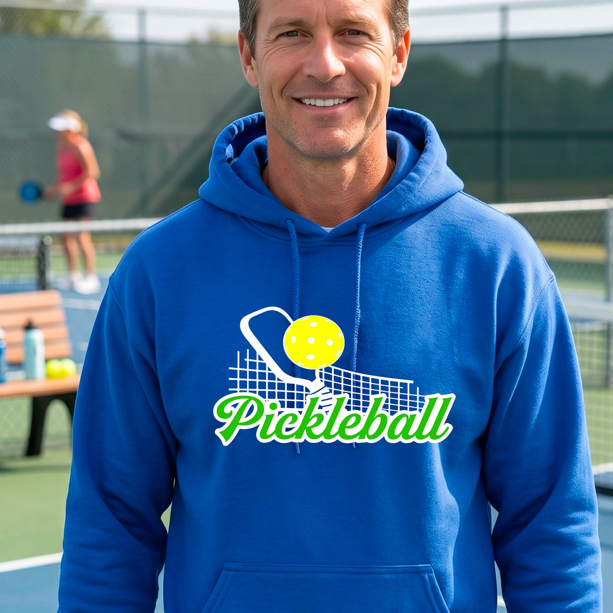 A close-up of a man wearing a royal blue long-sleeve unisex hoodie pullover with a graphic on the front that has white and green text "PICKLEBALL'  with a white pickleball paddle and net and a yellow pickleball above the text graphic.