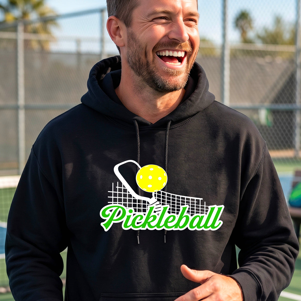 A close-up of a man wearing a black long-sleeve unisex hoodie pullover with a graphic on the front that has white and green text "PICKLEBALL'  with a white pickleball paddle and net and a yellow pickleball above the text graphic.