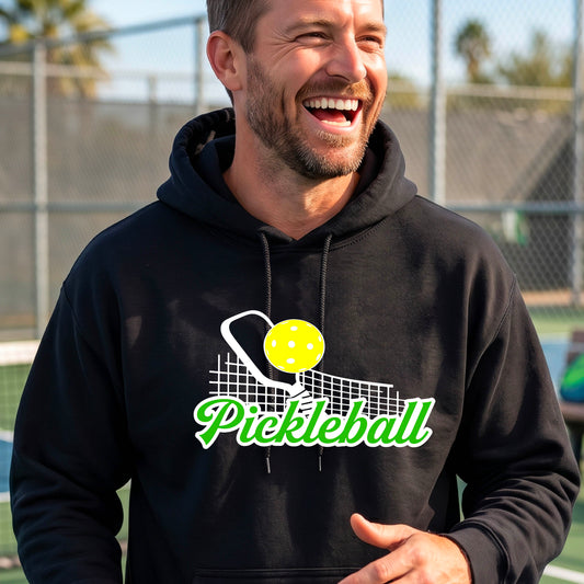 A close-up of a man wearing a black long-sleeve unisex hoodie pullover with a graphic on the front that has white and green text "PICKLEBALL'  with a white pickleball paddle and net and a yellow pickleball above the text graphic.