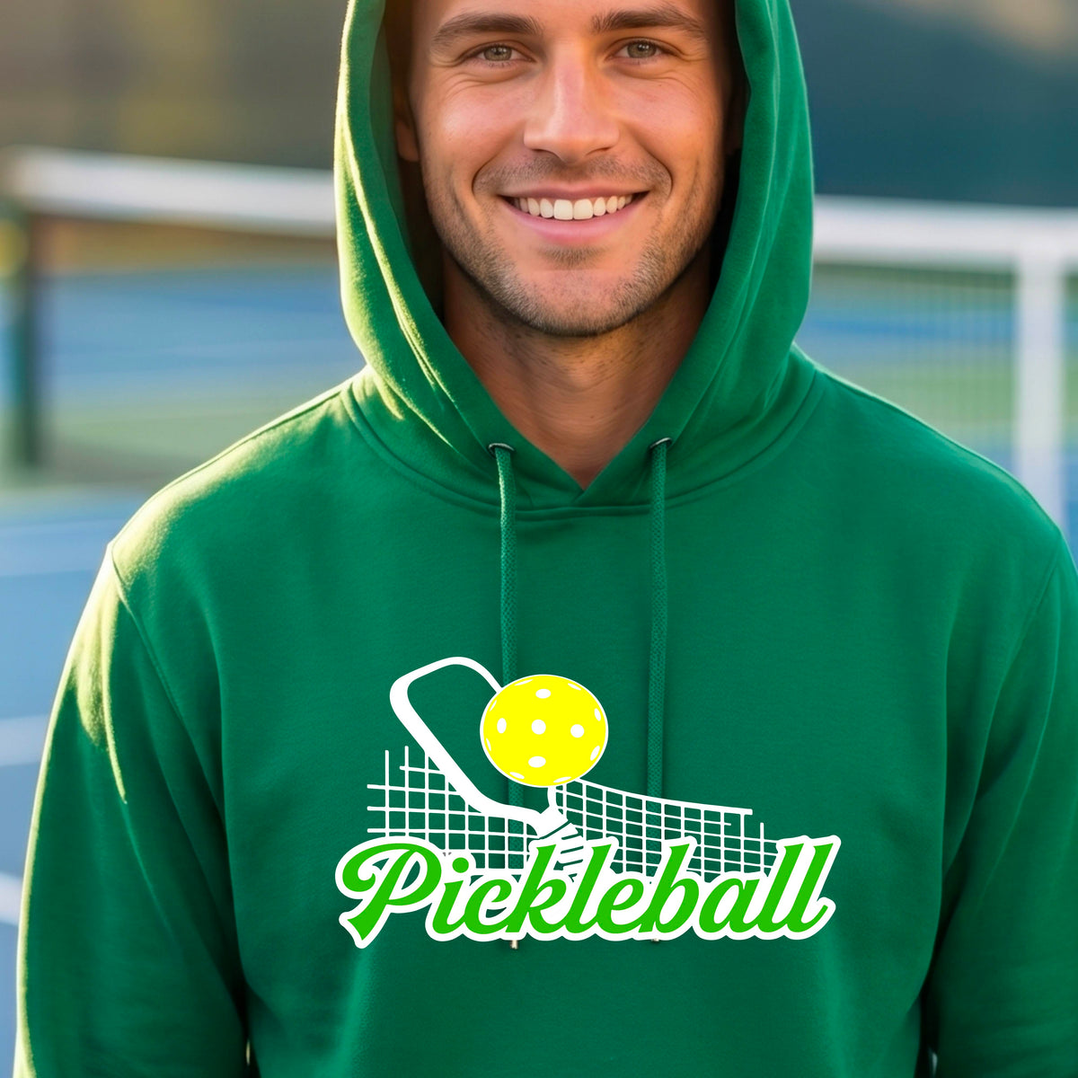 A close-up of a man wearing a green long-sleeve unisex hoodie pullover with a graphic on the front that has white and green text "PICKLEBALL'  with a white pickleball paddle and net and a yellow pickleball above the text graphic.