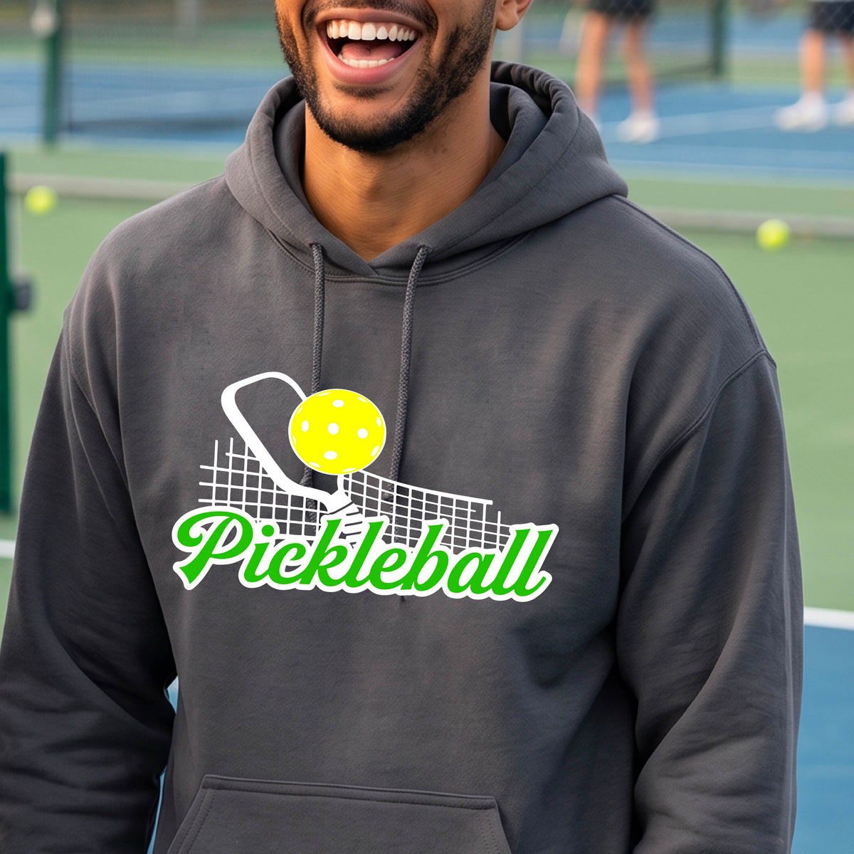 A close-up of a man wearing a gray long-sleeve unisex hoodie pullover with a graphic on the front that has white and green text "PICKLEBALL'  with a white pickleball paddle and net and a yellow pickleball above the text graphic.