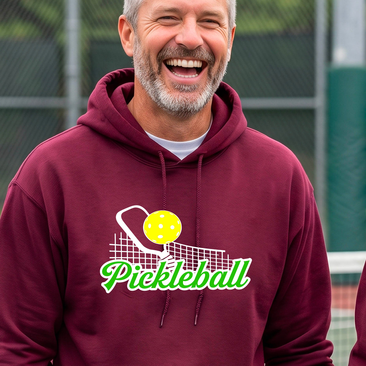 A close-up of a man wearing a maroon long-sleeve unisex hoodie pullover with a graphic on the front that has white and green text "PICKLEBALL'  with a white pickleball paddle and net and a yellow pickleball above the text graphic.