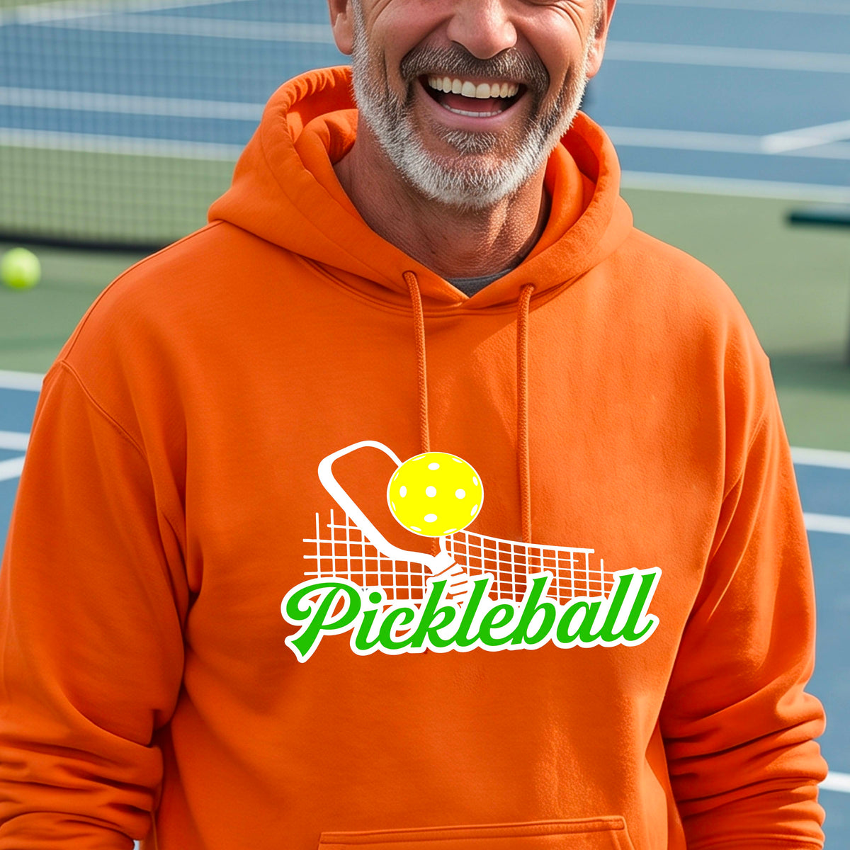 A close-up of a man wearing an orange long-sleeve unisex hoodie pullover with a graphic on the front that has white and green text "PICKLEBALL'  with a white pickleball paddle and net and a yellow pickleball above the text graphic.