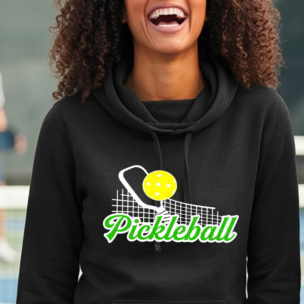 A close-up of a woman wearing a black long-sleeve cowl-neck hoodie pullover with a graphic on the front that has white and green text "PICKLEBALL'  with a white pickleball paddle and net and a yellow pickleball above the text graphic.