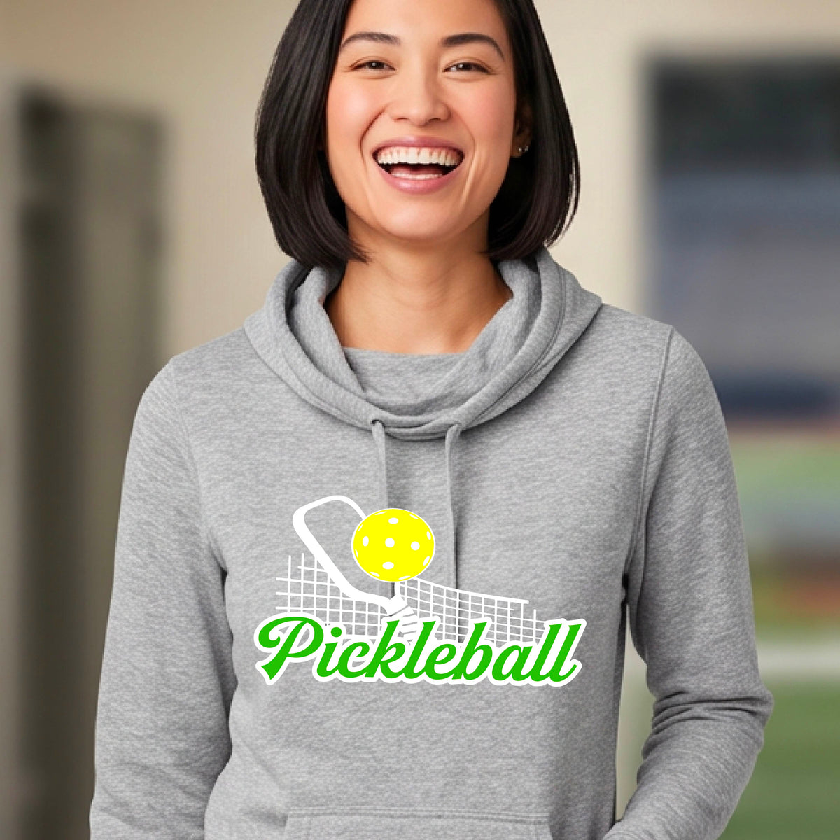 A close-up of a woman wearing a light gray long-sleeve cowl-neck hoodie pullover with a graphic on the front that has white and green text "PICKLEBALL'  with a white pickleball paddle and net and a yellow pickleball above the text graphic.