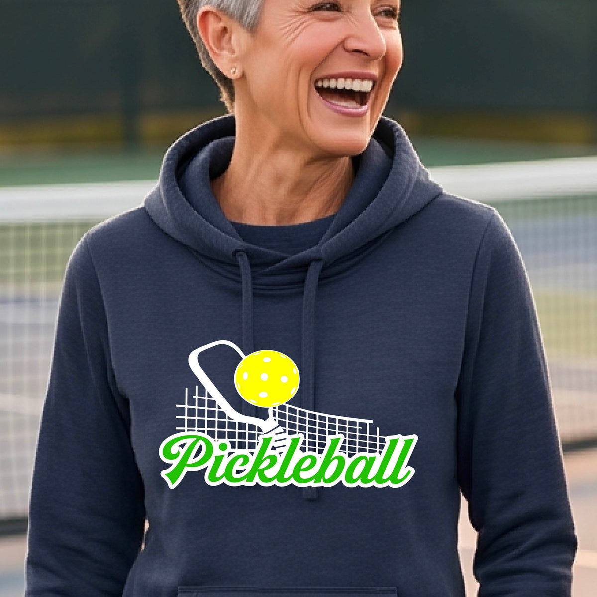 A close-up of a woman wearing a navy blue long-sleeve cowl-neck hoodie pullover with a graphic on the front that has white and green text "PICKLEBALL'  with a white pickleball paddle and net and a yellow pickleball above the text graphic.