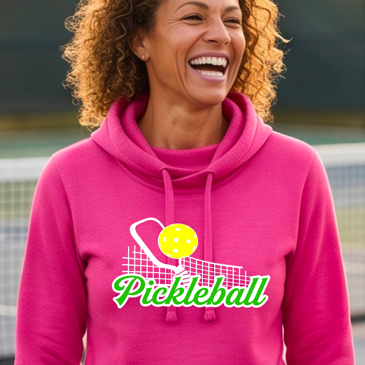A close-up of a woman wearing a pink long-sleeve cowl-neck hoodie pullover with a graphic on the front that has white and green text "PICKLEBALL'  with a white pickleball paddle and net and a yellow pickleball above the text graphic.