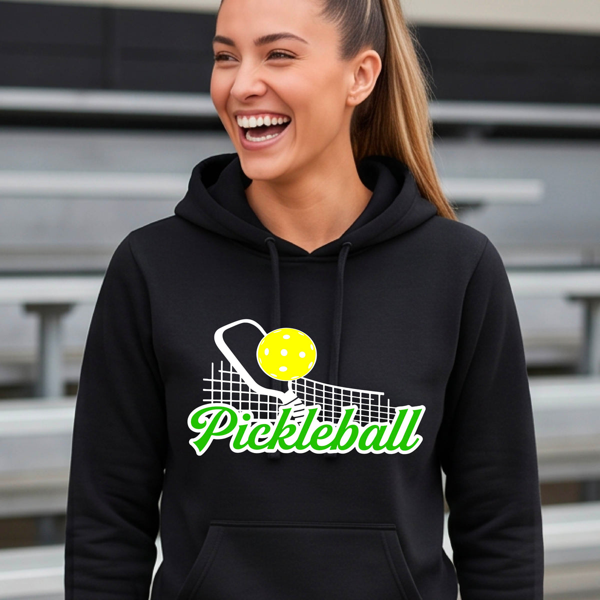 A close-up of a woman wearing a black long-sleeve fitted hoodie pullover with a graphic on the front that has white and green text "PICKLEBALL'  with a white pickleball paddle and net and a yellow pickleball above the text graphic.