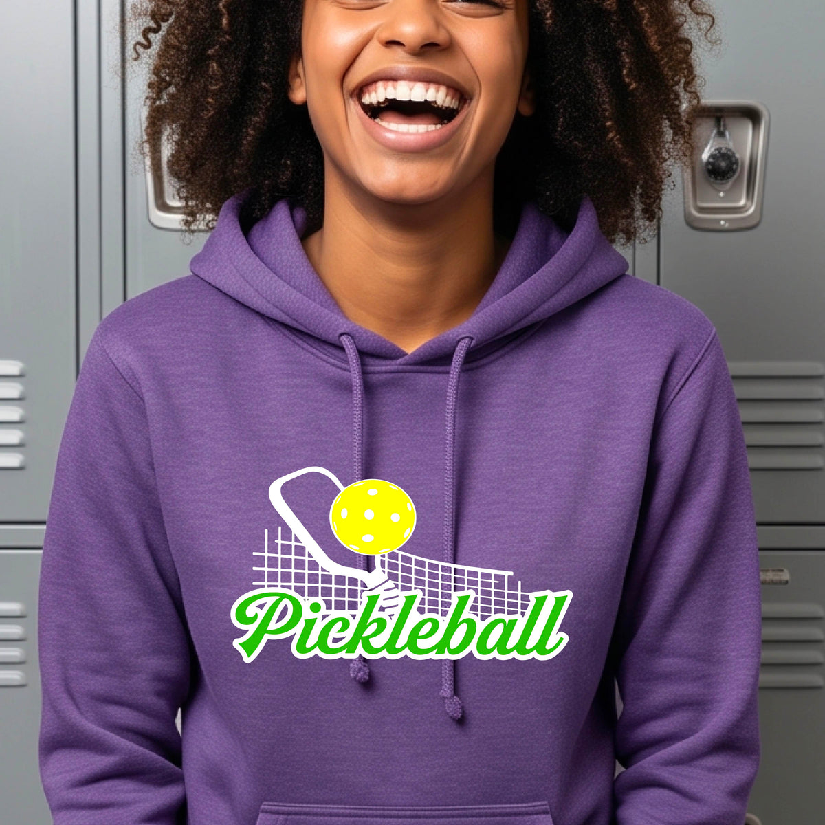 A close-up of a woman wearing a purple long-sleeve fitted hoodie pullover with a graphic on the front that has white and green text "PICKLEBALL'  with a white pickleball paddle and net and a yellow pickleball above the text graphic.