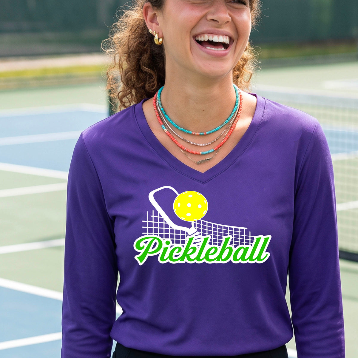 A woman wearing a purple long-sleeve v-neck t-shirt with a graphic on the front that has white and green text "PICKLEBALL'  with a white pickleball paddle and net and a yellow pickleball above the text graphic.