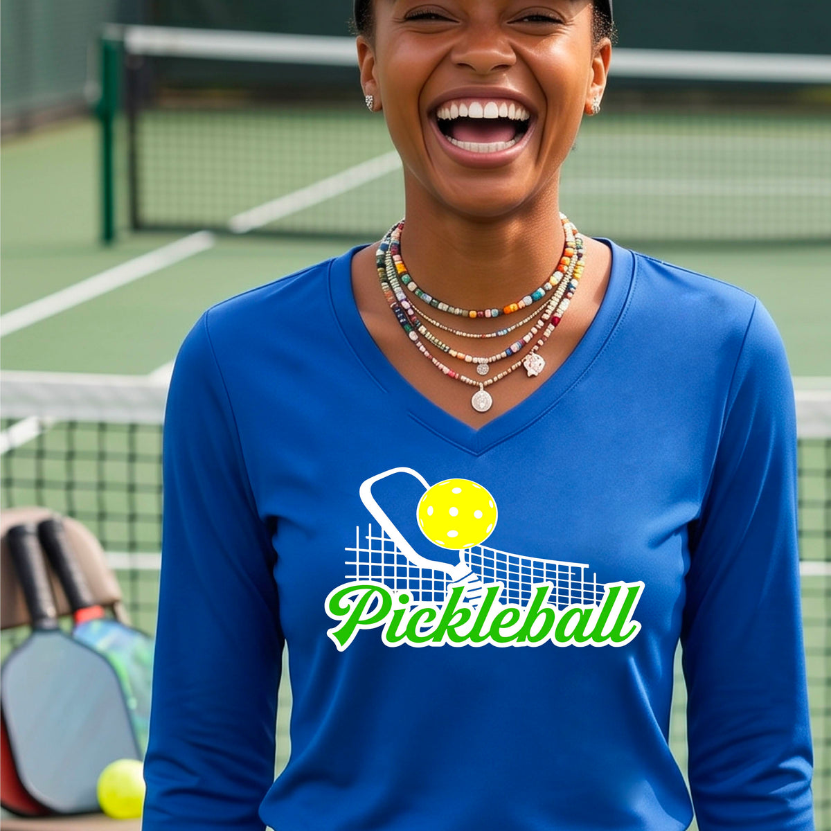 A woman wearing a royal blue long-sleeve v-neck t-shirt with a graphic on the front that has white and green text "PICKLEBALL'  with a white pickleball paddle and net and a yellow pickleball above the text graphic.