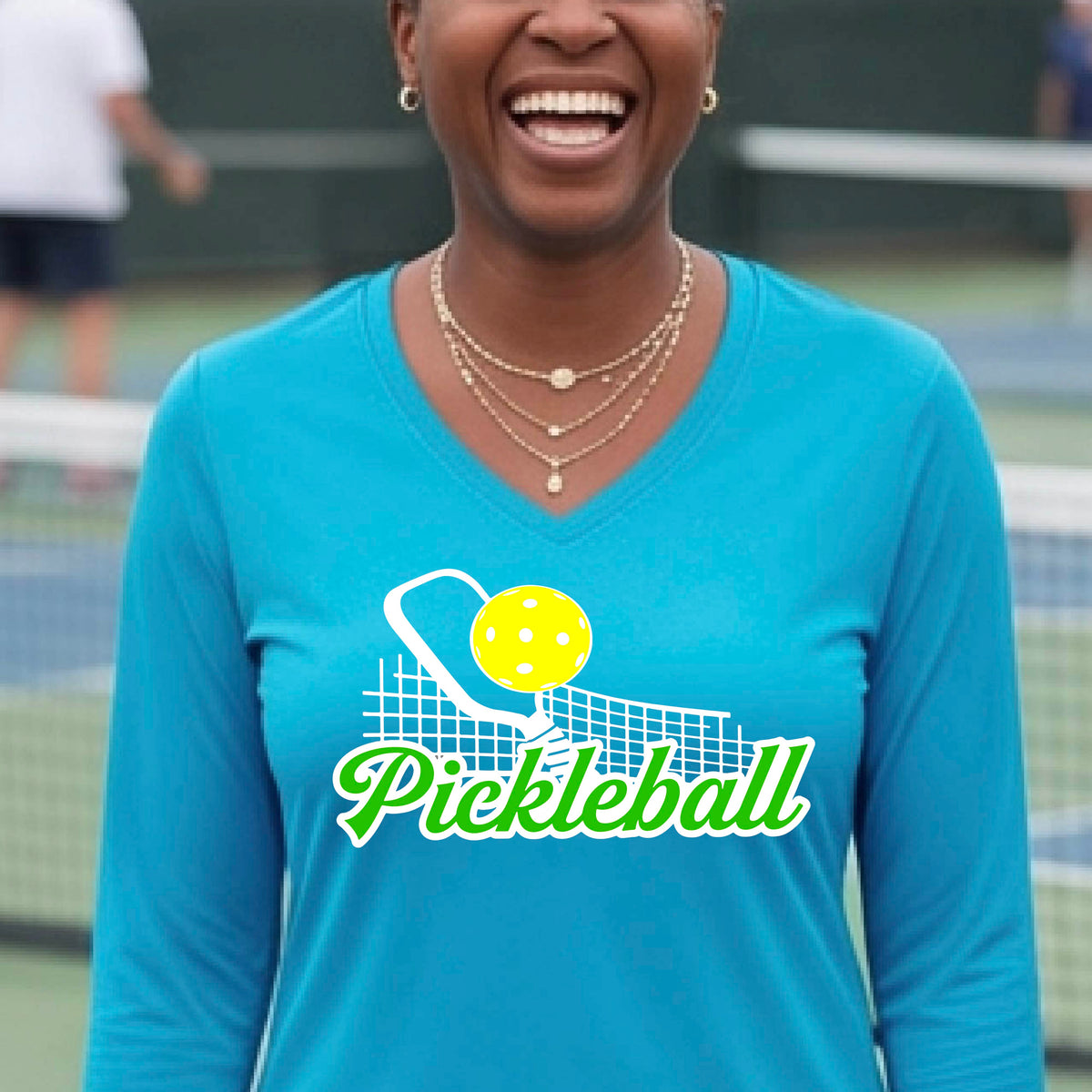 A woman wearing a bright bluelong-sleeve v-neck t-shirt with a graphic on the front that has white and green text "PICKLEBALL'  with a white pickleball paddle and net and a yellow pickleball above the text graphic.