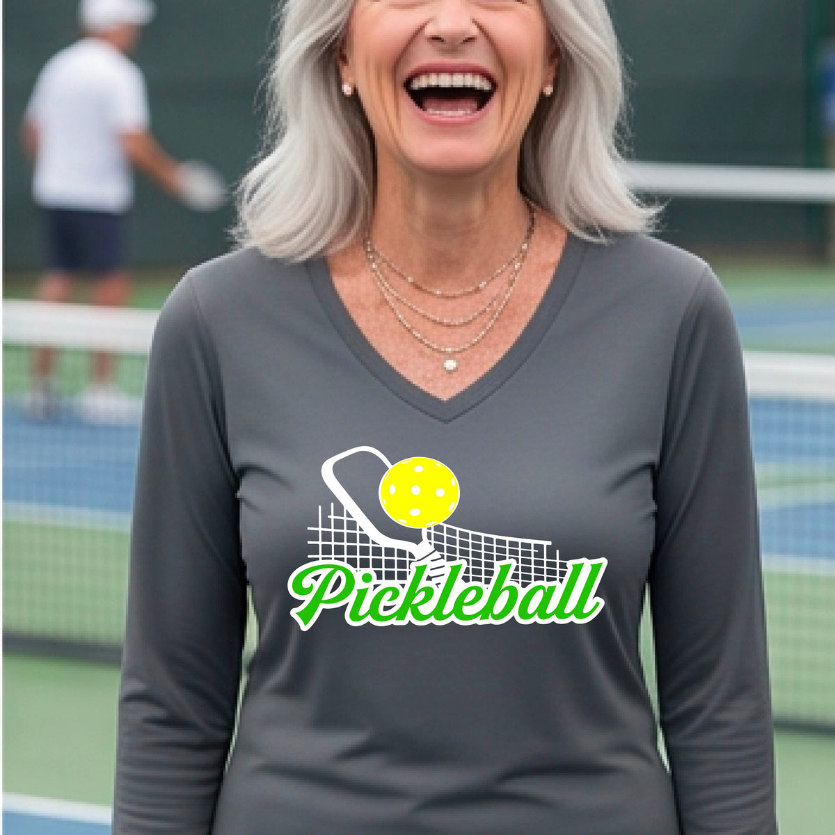 A woman wearing a gray long-sleeve v-neck t-shirt with a graphic on the front that has white and green text "PICKLEBALL'  with a white pickleball paddle and net and a yellow pickleball above the text graphic.