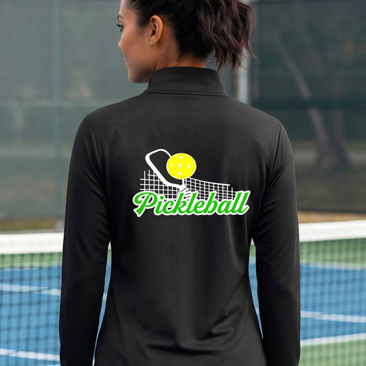 A woman viewed from behind wearing a black long-sleeve athletic qurter-zip jacket on a pickleball court. The back of the jacket features a graphic with a yellow pickleball, white net, and green 'Pickleball' text.