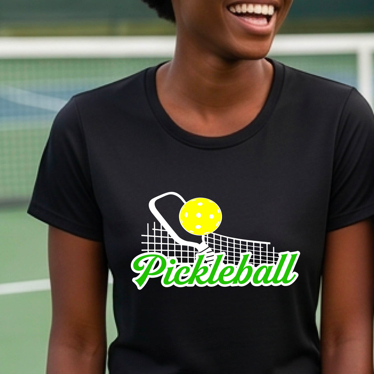 A woman wearing a black short-sleeve t-shirt with a graphic on the front that has white and green text "PICKLEBALL'  with a white pickleball paddle and net and a yellow pickleball above the text graphic.