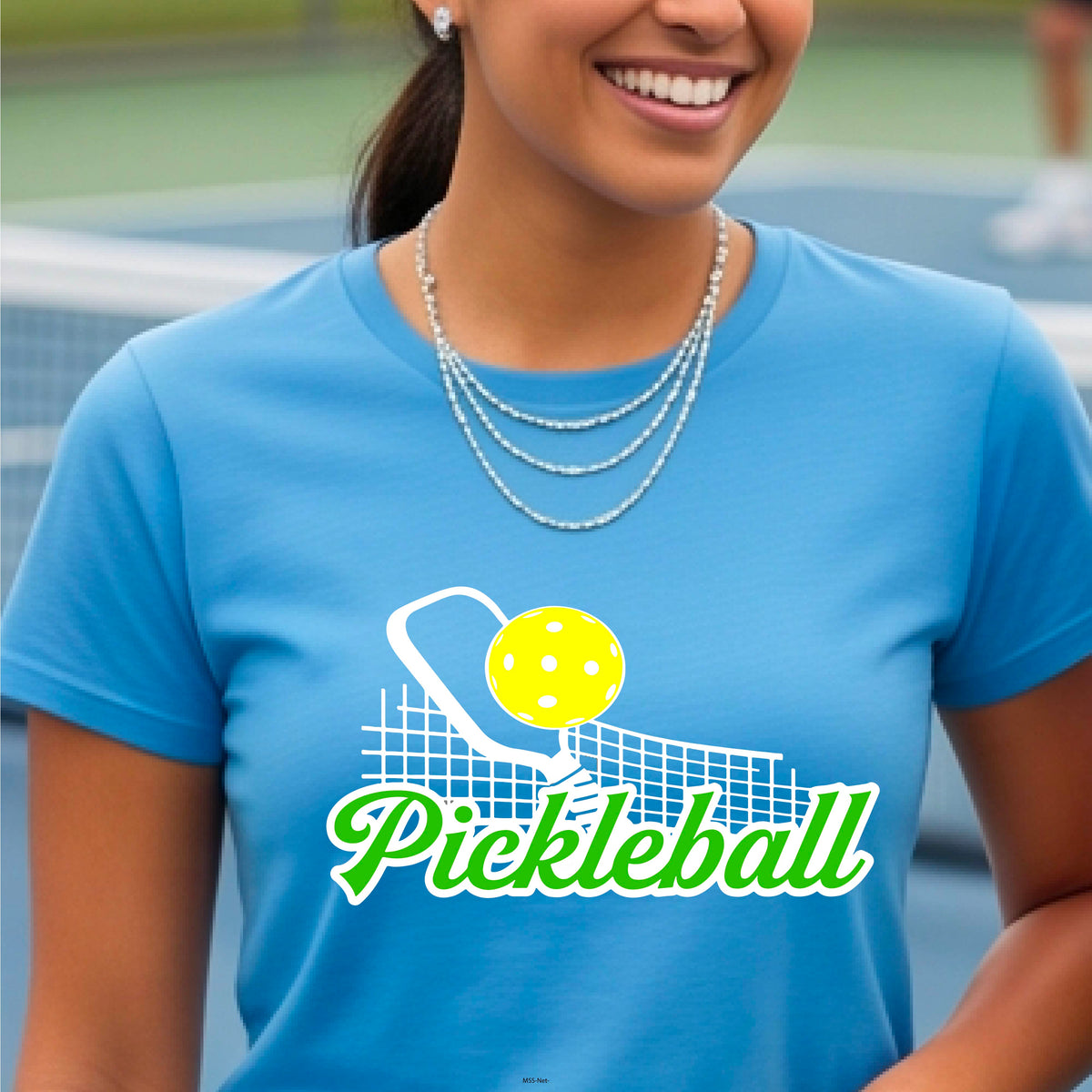 A woman wearing a bright blue short-sleeve t-shirt with a 'Pickleball' graphic on the front, featuring a yellow pickleball, white paddle, and green text, set against a blurred outdoor pickleball court background.