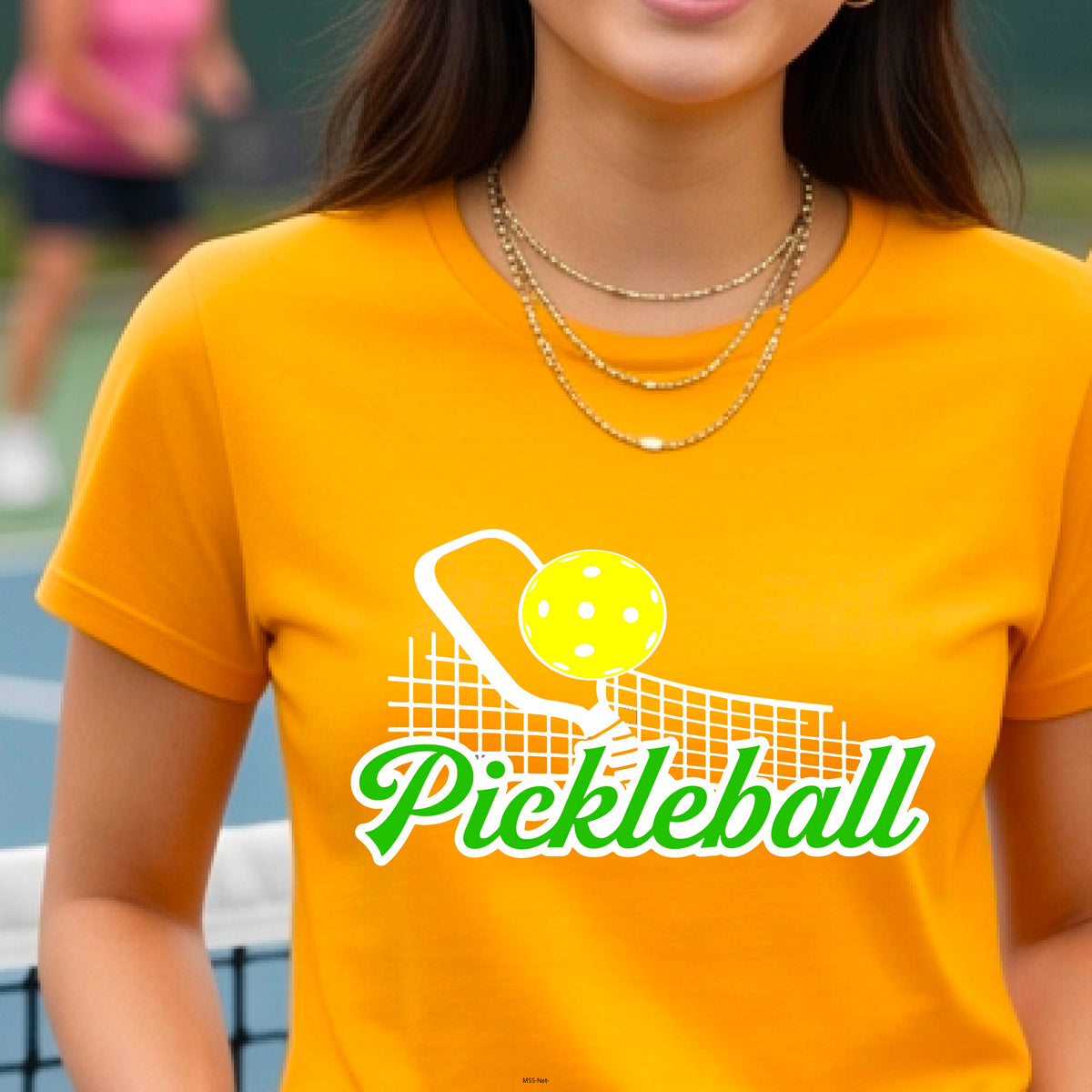 A woman wearing a mustard yellow gold short-sleeve t-shirt with a graphic on the front that has white and green text "PICKLEBALL'  with a white pickleball paddle and net and a yellow pickleball above the text graphic.