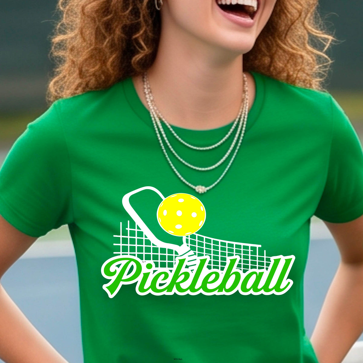 A woman wearing a green short-sleeve t-shirt with a graphic on the front that has white and green text "PICKLEBALL'  with a white pickleball paddle and net and a yellow pickleball above the text graphic.