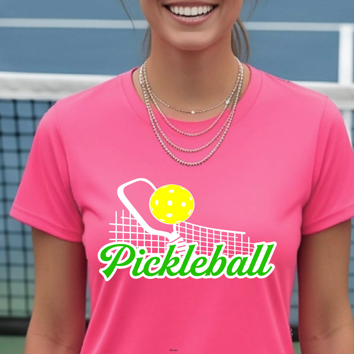 A woman wearing a bright pink short-sleeve t-shirt with a 'Pickleball' graphic on the chest, standing in front of a blue pickleball court net.