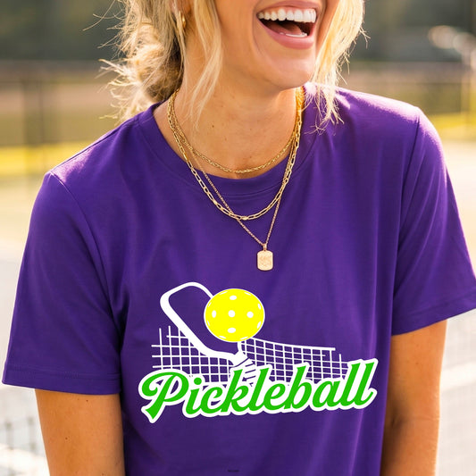 A woman wearing a purple short-sleeve t-shirt with a graphic on the front that has white and green text "PICKLEBALL'  with a white pickleball paddle and net and a yellow pickleball above the text graphic.