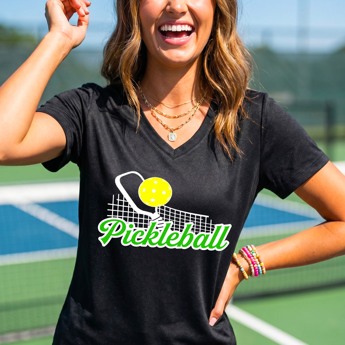 A woman wearing a black short-sleeve v-neck t-shirt with a graphic on the front that has white and green text "PICKLEBALL'  with a white pickleball paddle and net and a yellow pickleball above the text graphic.