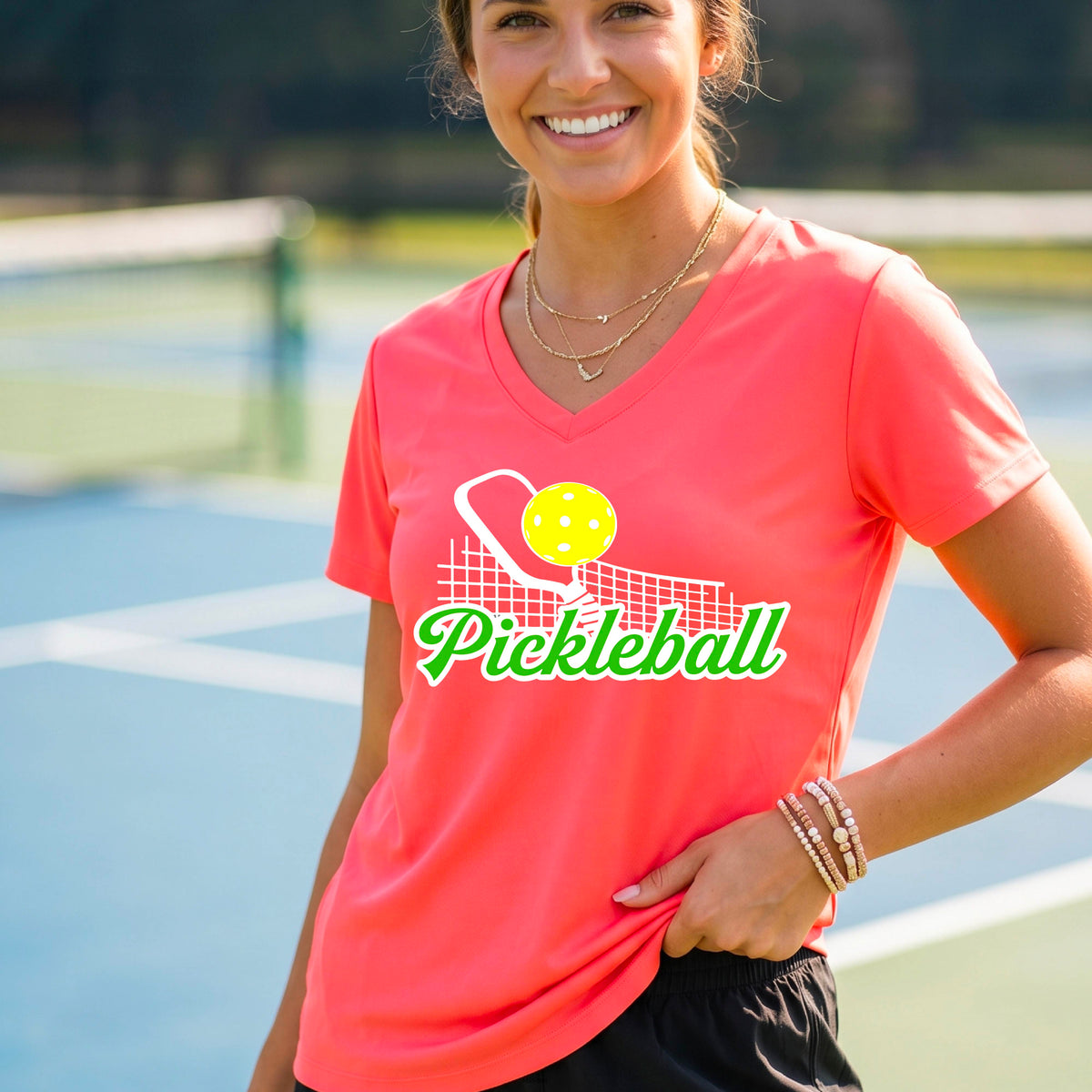 A woman on a pickleball court wearing a coral pink V-neck t-shirt with a graphic of a paddle, ball, and net above the word 'Pickleball' in green and white.