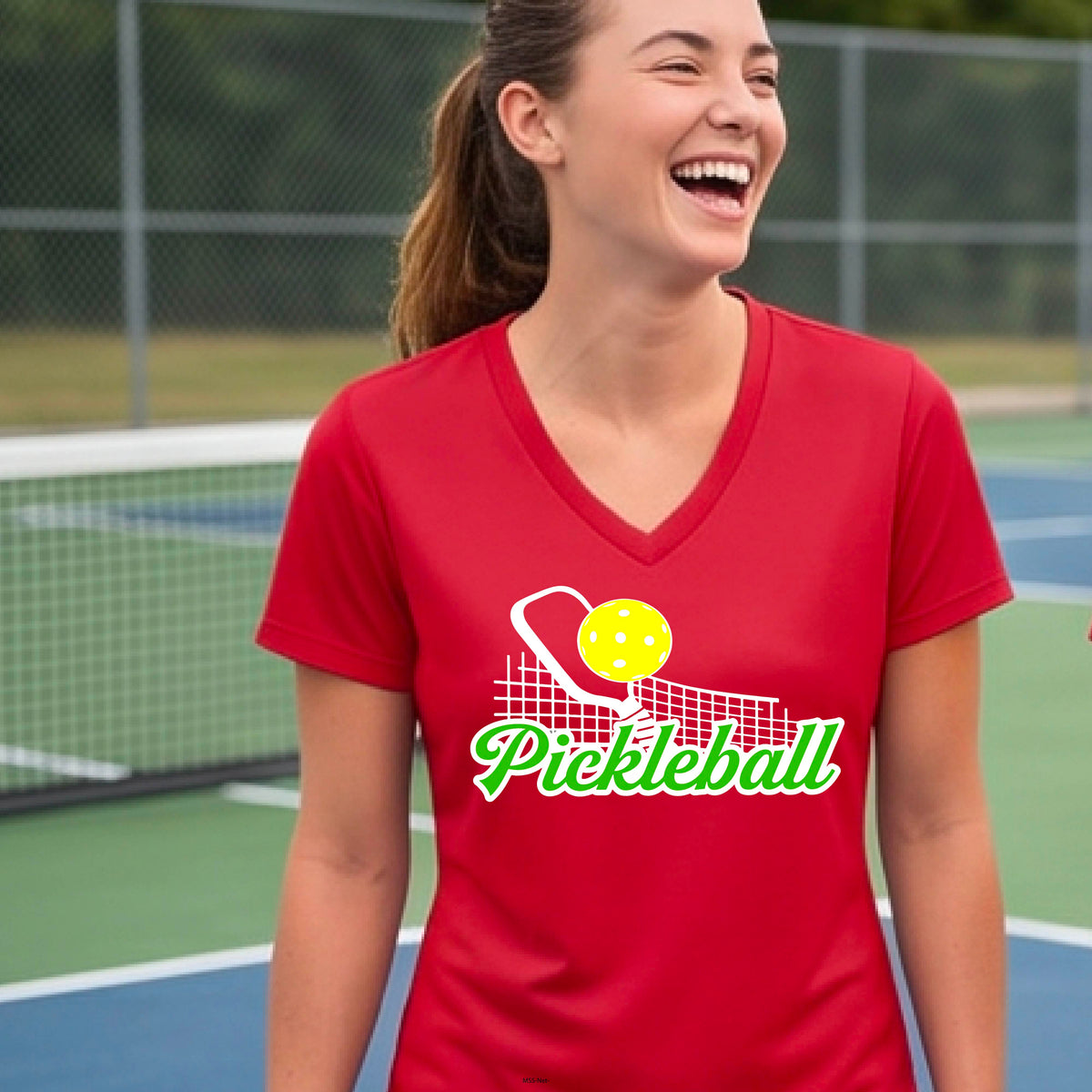 A woman laughing on a pickleball court wearing a red V-neck t-shirt with a yellow pickleball and green 'Pickleball' text graphic on the front.