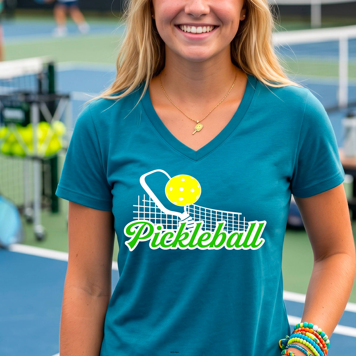 A woman wearing a teal short-sleeve v-neck t-shirt with a graphic on the front that has white and green text "PICKLEBALL'  with a white pickleball paddle and net and a yellow pickleball above the text graphic.