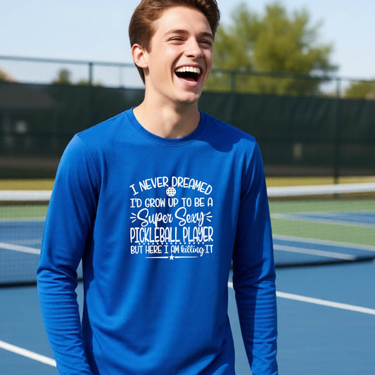 A young person on a pickleball court wearing a royal blue long-sleeve t-shirt with a white graphic of the text:  ‘I Never Dreamed I’d Grow Up To Be A Super Sexy Pickleball Player But Here I Am Killing It!’, standing on a pickleball court.