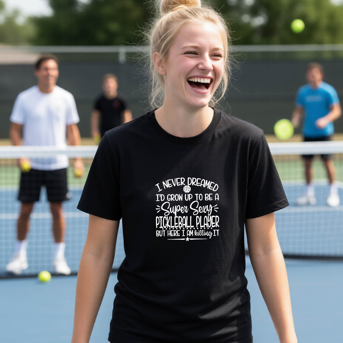 A young person on a pickleball court wearing a black long-sleeve t-shirt with a white graphic of the text:  ‘I Never Dreamed I’d Grow Up To Be A Super Sexy Pickleball Player But Here I Am Killing It!’, standing on a pickleball court.