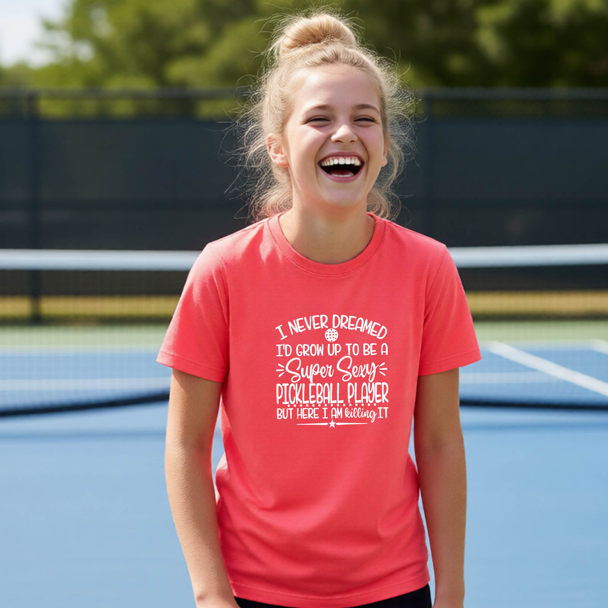 A young person on a pickleball court wearing a coral pink long-sleeve t-shirt with a white graphic of the text:  ‘I Never Dreamed I’d Grow Up To Be A Super Sexy Pickleball Player But Here I Am Killing It!’, standing on a pickleball court.