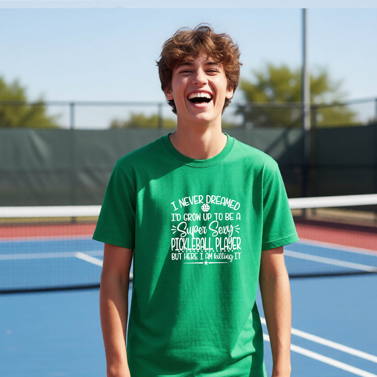 A young person on a pickleball court wearing a green long-sleeve t-shirt with a white graphic of the text:  ‘I Never Dreamed I’d Grow Up To Be A Super Sexy Pickleball Player But Here I Am Killing It!’, standing on a pickleball court.
